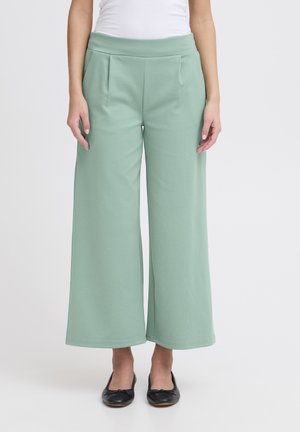 Pantalons larges vert clair en tissu doux, dotés d'une taille haute et de plis pour ajouter de la texture. Associés à des ballerines noires.