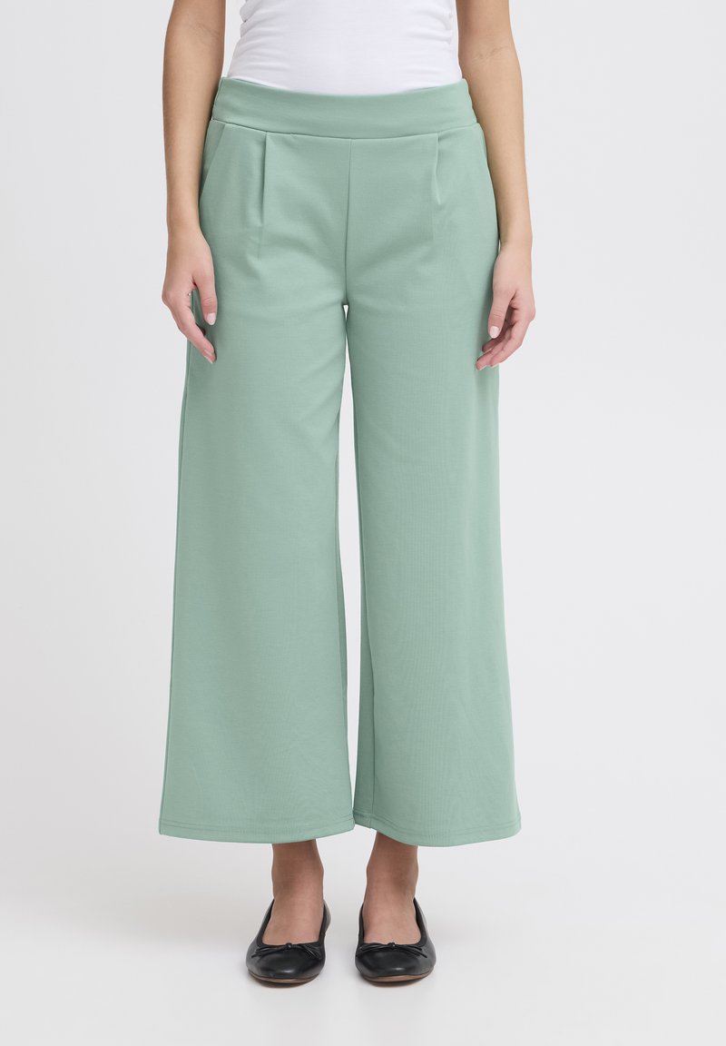 Lichtgroene wijde broek van zachte stof, met een hoge tailleband en plooien voor extra textuur. Gepaarde met zwarte platte schoenen.