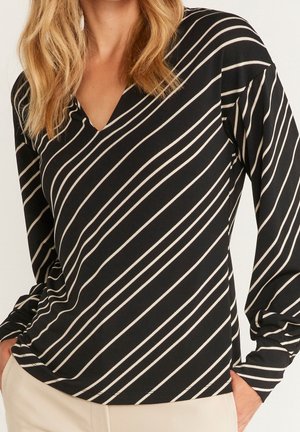 Femme portant un haut noir à manches longues avec des rayures diagonales blanches et un col en V, mains dans les poches, pantalon beige visible à la taille.