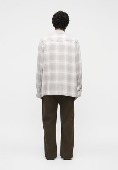 Άτομο με σγουρά μαλλιά, γυρισμένο με την πλάτη, που φορά ανοιχτό γκρι καρό μακρυμάνικο πουκάμισο και άνετο σκούρο καφέ παντελόνι μπροστά σε μονόχρωμο φόντο.