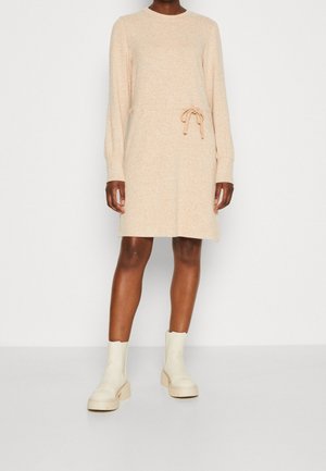 Robe beige en tricot avec un col rond, des manches longues et un cordon de serrage à la taille. Portée avec des bottines crème.