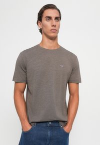 Brun kortermet T-skjorte laget av mykt stoff med rund hals. Har en liten brodert logo på brystet. Stilt med blå jeans.