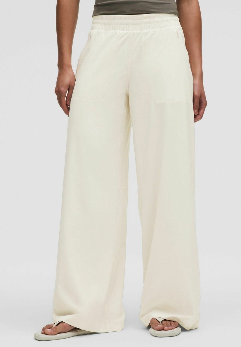 Personne portant un pantalon large ample de couleur blanc cassé avec taille élastique et des sandales tongs, debout devant un fond uni.