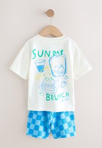 Chemise blanche à manches courtes avec "Sunday Brunch Club" et dessins de petit-déjeuner, associée à un short bleu à carreaux suspendu sur un cintre en bois.