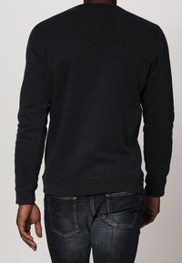 Pullover nero con colletto tondo e maniche lunghe, realizzato in tessuto morbido. Presenta polsini e orlo a coste, indossato con jeans di denim blu.