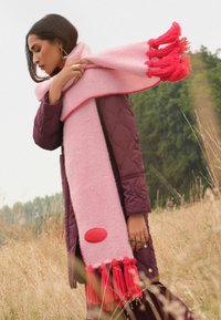 Długi różowy wełniany szalik z frędzlami na końcach w intensywnym różu, z gładkim czerwonym akcentem i kontrastującymi czerwonymi przeszyciami wzdłuż brzegów.