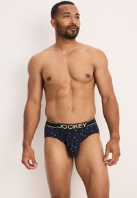 Slip da uomo navy con un motivo geometrico in verde e blu. Vita elastica con il nome del marchio "JOCKEY" in giallo. Materiale morbido.