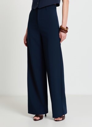 Femme portant un pantalon large bleu marine avec des talons ouverts noirs et des bracelets en bois marron au poignet gauche.