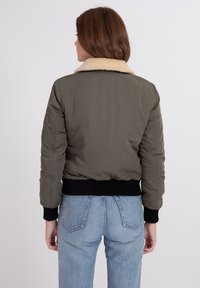 Giacca bomber verde oliva con colletto in pelliccia sintetica beige e polsini e orlo a righe neri. Presenta una finitura liscia e lucida e una silhouette aderente.