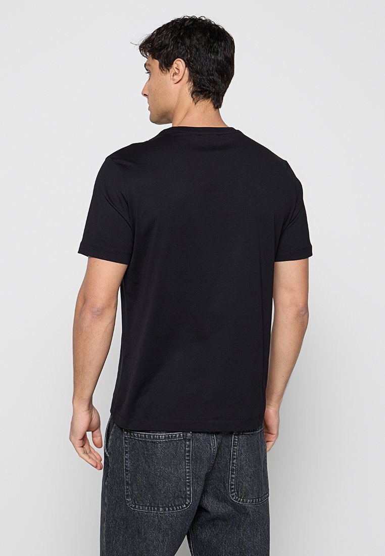 Hombre con cabello corto y oscuro, vistiendo una camiseta negra lisa de manga corta y vaqueros gris oscuro, de espaldas sobre un fondo blanco.
