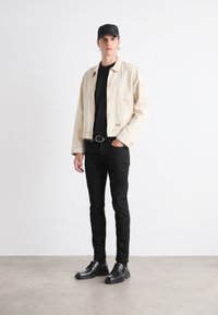Chaqueta de mezclilla beige con cierre de cremallera, camiseta negra, jeans skinny negros y zapatos de cuero negros. Hombre de pie contra un fondo neutro.