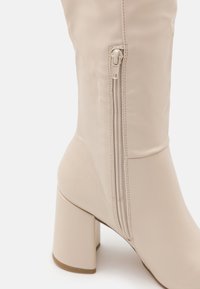 Beige knähöga stövlar i slät material, med sidodragkedja, kraftig klack och minimalistisch design med sömdetaljer.