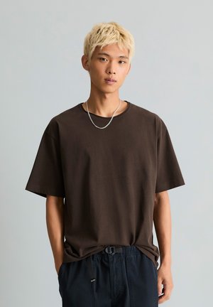 Młoda osoba o krótkich blond włosach, ubrana w luźny ciemnobrązowy t-shirt, srebrny łańcuszek i ciemne spodnie, z rękami w kieszeniach, neutralne tło.