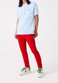 Ljusblå pikétröja med en grön logotyp, kombinerad med röda smala byxor och vita sneakers med gröna accenter och svarta detaljer.