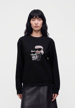 Mulher com cabelo preto comprido veste sweatshirt preta com desenho de strass de uma pessoa e um cão, ambos a usar óculos de sol.