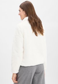 Witte fleece jacket met een hoge kraag en lange mouwen, met een zachte textuur. Het model draagt grijze broeken, gezien van achteren.