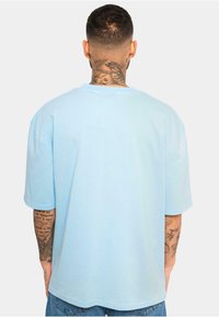 Tricou scurt cu mânecă scurtă de culoare albastru deschis, cu guler rotund și croială lejeră, fabricat din bumbac moale. Tatuaje vizibile pe antebrațe.