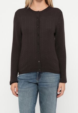 Femme portant un cardigan côtelé marron foncé boutonné et un jean bleu, debout devant un fond blanc uni.