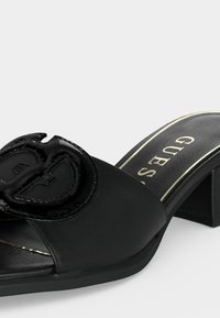 Μαύρο mule σανδάλι με ανοιχτό δάχτυλο και τετράγωνο τακούνι, με γυαλιστερό έμβλημα λογότυπου Guess στο φαρδύ μπροστινό λουράκι και χρυσή επένδυση στην εσωτερική σόλα.