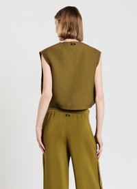 Olivgrünes ärmelloses Oberteil mit hohem Kragen und spitz zulaufendem Rücken, kombiniert mit weit geschnittenen Hosen mit seitlichen Knopfdetails.