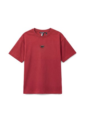 Camiseta roja de manga corta con cuello redondo y un pequeño parche negro y rojo centrado en el pecho.