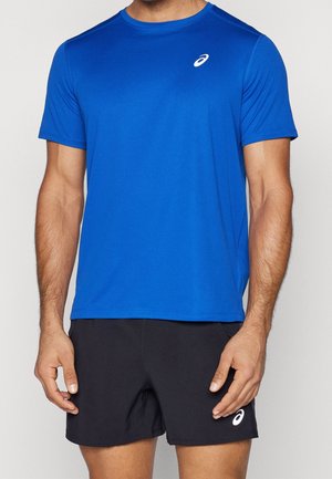 Mężczyzna w niebieskim sportowym t-shircie z krótkim rękawem i czarnych krótkich spodniach, oba z białymi logo ASICS, stoi na szarym tle.