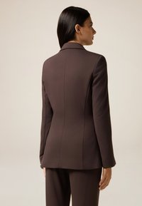 Blazer marron ajusté avec une silhouette cintrée, texture lisse, revers en pointe et manches longues. Les coutures soulignent la structure.