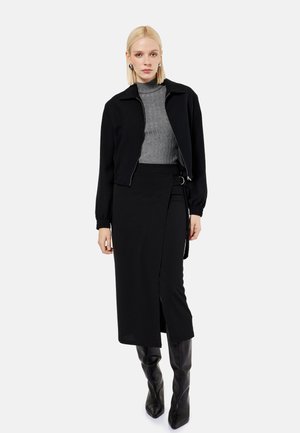 Vrouw draagt een zwarte cropped jas over een grijze geribde coltrui, een zwarte wikkelrok met split en zwarte kniehoge laarzen, staande op een witte achtergrond.