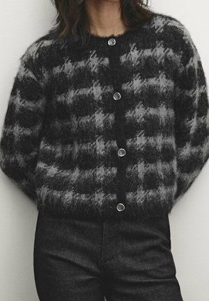 Femme portant un cardigan boutonné à motifs duveteux noirs et gris et un pantalon noir, debout devant un fond clair uni.