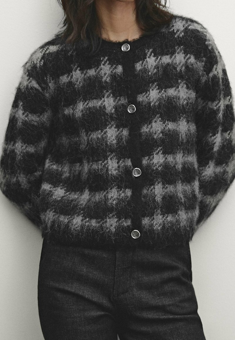 Femme portant un cardigan boutonné à motifs duveteux noirs et gris et un pantalon noir, debout devant un fond clair uni.