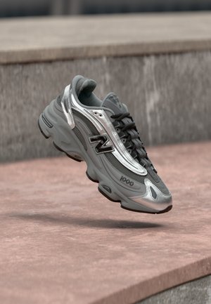 Baskets New Balance grises et argentées marquées "1000", flottant au-dessus d'une surface en pierre rosée avec un mur en pierre grise en arrière-plan.