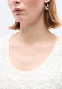 Λευκό τοπ με στρωματοποιημένη υφή από παγιέτες και ευρύ ντεκολτέ. Ασημένια σκουλαρίκια κρίκοι σε σχήμα δακρύου. Ο τόνος του δέρματος είναι ανοιχ τός.