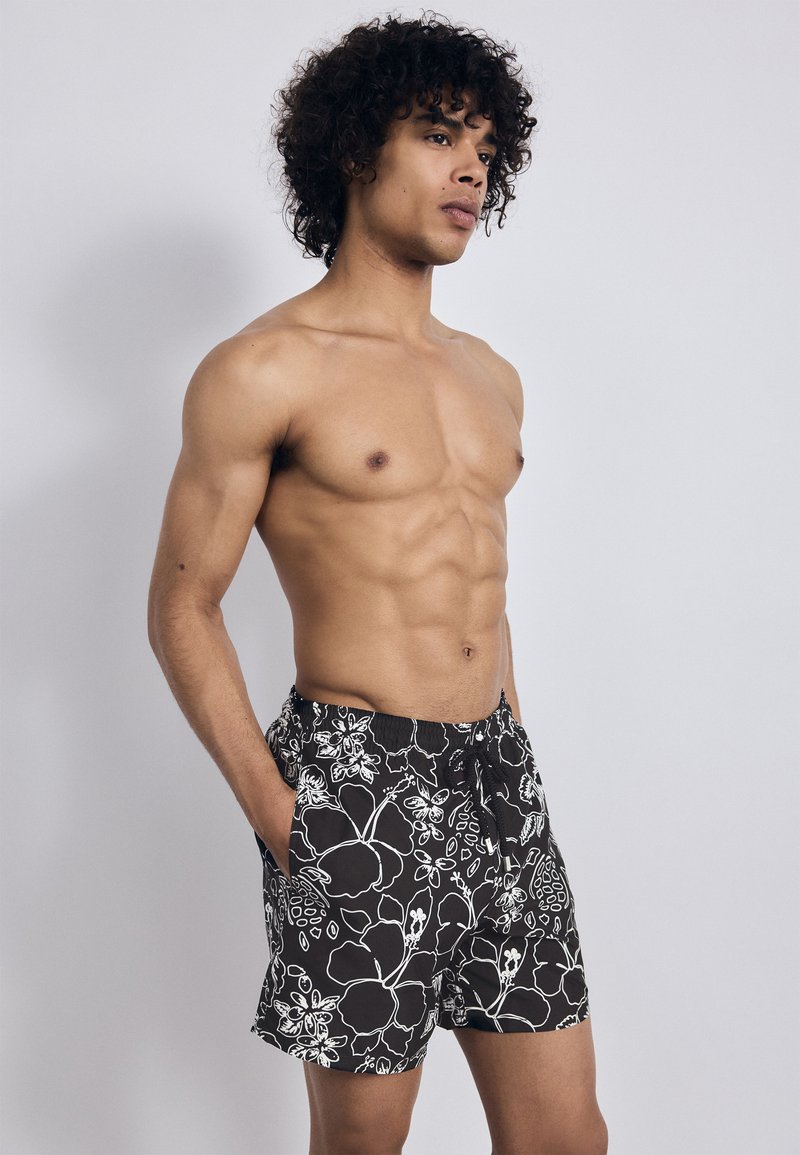 Muž bez trička s kučeravými vlasmi, oblečený v čiernych plavkách so bielym kvetinovým vzorom, stojí s rukami v vreckách a pozerá sa nabok.