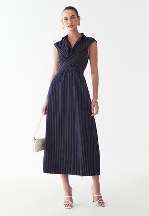 Marineblå stribet maxi-kjole med krave, korte ærmer, omslagdesgin og tætsiddende talje. Modellen bærer en lys taske og har hæle på.