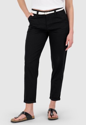 Frau trägt schwarze hochgekrempelte Hose, weißen geflochtenen Gürtel, weißes Oberteil und schwarze Sandalen mit Korksohle, steht mit einer Hand in der Hosentasche.