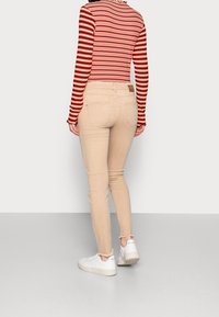 Mujer vistiendo jeans skinny beige, zapatillas blancas y un top de manga larga a rayas en rojo, rosa y negro, de pie contra un fondo claro.