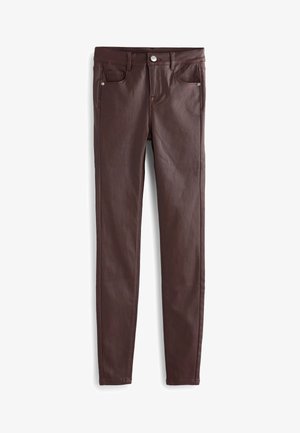 Mujer con top de punto sin mangas beige, pantalones pitillo de cuero sintético marrón oscuro y sandalias con tiras trenzadas beige sobre un fondo rosa pálido.