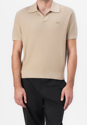 Hombre con polo de punto beige de manga corta y cuello, y pantalones negros, de pie frente a un fondo blanco liso.