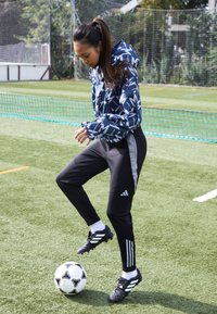 Sport jacket with navy and white geometric pattern, black athletic pants with gray side accents, soccer ball on artificial grass.