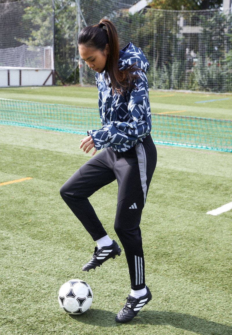 Sport jacket with navy and white geometric pattern, black athletic pants with gray side accents, soccer ball on artificial grass.