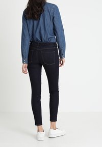 Jeans oscuros de corte slim con cintura alta, combinados con una camisa de mezclilla. La camisa tiene puños con botones y un detalle en la parte trasera. Zapatillas blancas completan el look.