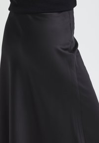 Jupe midi en satin noir portée avec un haut noir, mettant en valeur le tissu lisse et de légers détails de couture latérale.