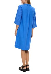 Robe chemise en lin bleu avec des manches 3/4, un col pointu et un détail au dos ; accompagnée de sandales à plateforme noires.