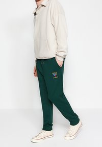 Beige mikina s límcem a přední kapsou, kombinovaná se zelenými tepláky s vyšitým logem, obutá s krémovými teniskami.