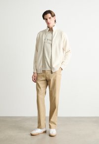 Krämfärgad zip-up-jacka över en ljusgrå t-shirt med grafik, tillsammans med beige byxor och vita sneakers med beige accenter.