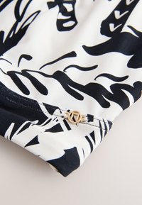 Tissu à motifs noir et blanc avec des formes abstraites et un petit logo métallique circulaire doré fixé près du bord.
