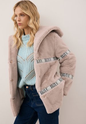 Jonge vrouw met golvend blond haar die een lichtblauwe gebreide trui en een beige zachte jas met BIZUU-logo banden draagt, staand tegen een bleke achtergrond.