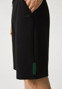 Shorts noirs en tissu doux avec poches latérales, arborant une étiquette texturée verte et un logo noir sur le devant.