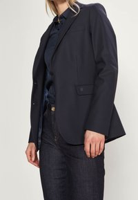 Läskare blazer av marinblått skräddarsytt tyg med två framknappar, en bröstficka och slagkragar. Kombineras med mörka jeans.