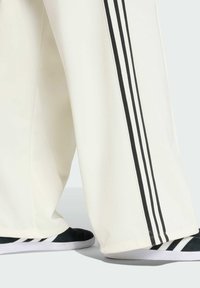 Bijele atletske hlače s crnim trostrukim detaljima na stranama, u paru s crno-bijelim atletskim tenisicama. Tkanina ima teksturirani završetak.